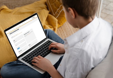 Ein Junge sitzt auf einem Sofa mit einem gelben Kissen und verwendet ein Laptop. Auf dem Bildschirm wird eine Online-Schulplattform angezeigt.